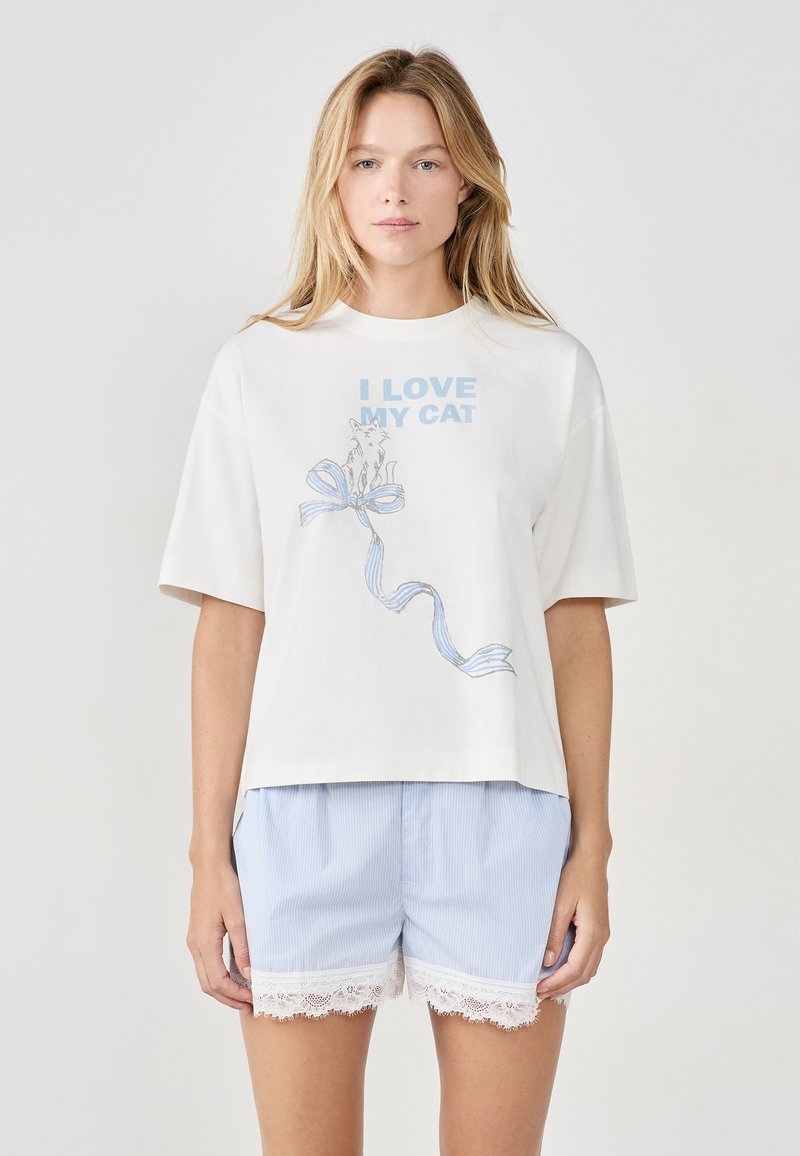Mujer rubia vistiendo una camiseta blanca con el texto "AMO A MI GATO" y una ilustración de un gato, combinada con pantalones cortos a rayas azul claro con encaje.
