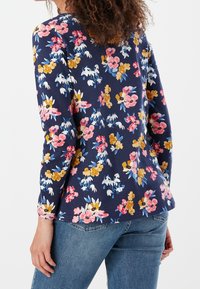 Květinová blůza s dlouhými rukávy v námořnické látce, zdobená růžovými, žlutými a modrými květinami. Obroubený lem a volný střih. Spárována s denimovými džíny.