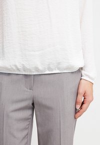 Personne portant un pantalon gris clair sur mesure et un haut blanc texturé à manches longues, ample, avec ourlet et poignets élastiqués.