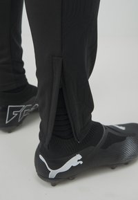 Black athletic pants with textured pattern, features zippered cuffs. Paired with black soccer cleats, displaying a white Puma logo.