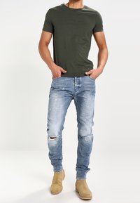 Muž oblečený v tmavě zelené košili s krátkým rukávem, světlých slim jeansových kalhotách se třenicemi a béžových semišových botách, stojí s rukama v kapsách na jednobarevném pozadí.