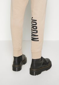 Beige Jogginghose mit einem großen schwarzen "JORDAN"-Logo an der Seite, kombiniert mit glänzenden schwarzen Knöchelstiefeln mit dicken Sohlen.