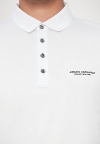 Polo shirt blanc avec col classique, comportant cinq boutons noirs et un logo en relief sur le côté gauche en texte noir : "ARMANI EXCHANGE MILANO/NEW YORK."