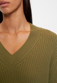 Žena v olivově zeleném ribbed pleteném svetru s výstřihem do V, odhalující krk, klíční kost a část obličeje s dlouhými tmavými vlasy.