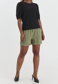 Kvinna iklädd en svart texturerad kortärmad topp, olivgröna shorts med dragsko och svarta spetsiga högklackade skor som står mot vit bakgrund.