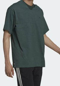 Ein Mann trägt ein dunkelgrünes T-Shirt mit kurzen Ärmeln und schwarze Hosen mit weißen Streifen an den Seiten und steht vor einem einfarbigen Hintergrund.