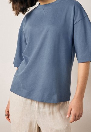 Osoba ubrana w luźną, zakurzononiebieską koszulę z krótkim rękawem i jasnobeżowe lniane spodnie stojąca na jednolitym tle.