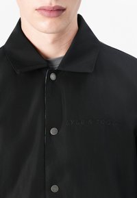 Veste légère noire avec col, ornée de boutons-pression en métal argenté et d'un logo en relief sur le côté gauche. Texture lisse et design minimaliste.