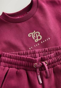 Maroninvärinen huppari, jossa on ribbauskaulus, ja jota koristi kultainen "TB"-logo sekä nyörikoriste. Pehmeä tekstuuri, kerätty vyötärö säilyttää säädettävän istuvuuden.