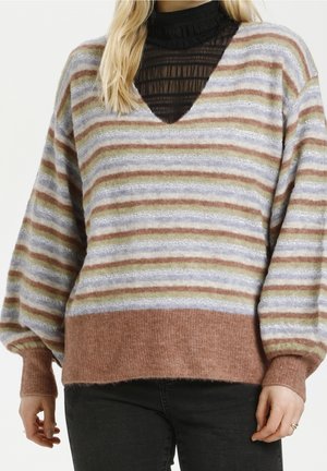 Mujer con un suéter de punto multicolor a rayas, con mangas farol y puños acanalados, sobre una camiseta negra transparente de cuello alto.