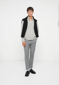 Conjunto masculino que presenta un chaleco de punto negro sobre una camisa de botones a rayas gris claro, pantalones grises de corte ajustado y mocasines negros.
