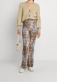 Cárdigan de punto en beige con tres botones, combinado con pantalones acampanados estampados en tonos tierra. Calzado: zapatillas claras, llevando una bolsa pequeña.