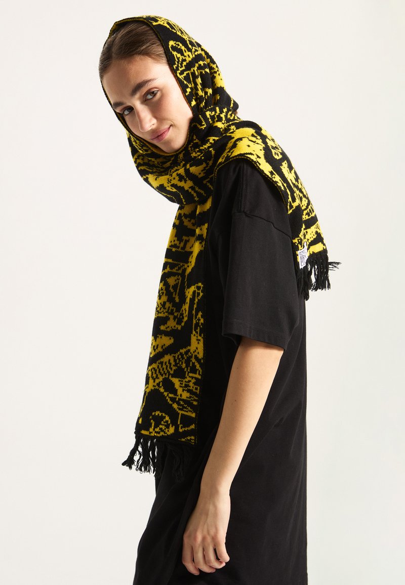 Sciarpa con motivo giallo e nero, con frange, indossata come cappuccio. Realizzata in morbido materiale lavorato a maglia, ricade su una maglietta nera. Dettagli di design intricate visibili.