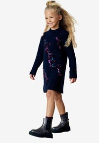 Robe pull en laine polaire bleu marine avec des manches longues, ornée de détails en sequins colorés. Assortie à des bottines grises de cheville avec des semelles épaisses.