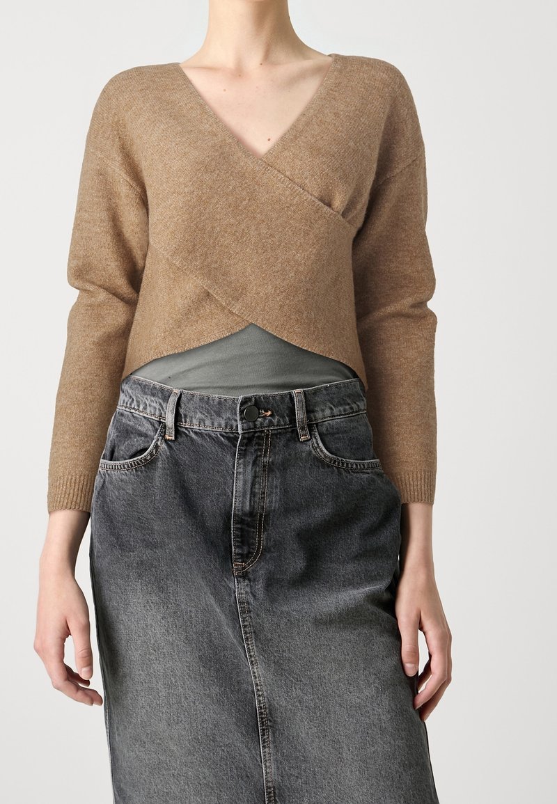 Donna che indossa un maglione avvolgente marrone a scollo a V sopra una maglietta grigia, abbinato a una gonna di jeans nera a vita alta, in piedi davanti a uno sfondo neutro.