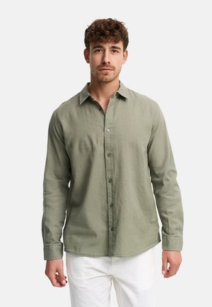 Młody mężczyzna o kręconych brązowych włosach, ubrany w oliwkowo-zieloną koszulę na guziki i białe spodnie, stojący na tle jednolitego jasnego tła.