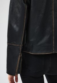 Giubbotto in pelle nera con finitura strutturata, dotato di cuciture marroni e dettaglio con zip sui polsini, visto da dietro.