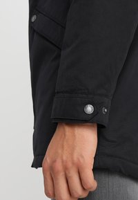 Manga de chaqueta negra con un acabado texturizado, que presenta un cierre de botón y un detalle de logo. El puño está ligeramente enrollado para revelar la muñeca.