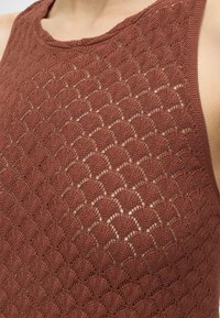 Gros plan du haut du torse portant un haut sans manches en tricot avec un motif en dentelle brune festonnée.