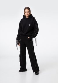 Czarny komplet z kapturem i spodniami, z ozdobnymi sznurowaniami na rękawach i bokach. Gładki materiał, luźny krój i okrągłe logo na bluzie.