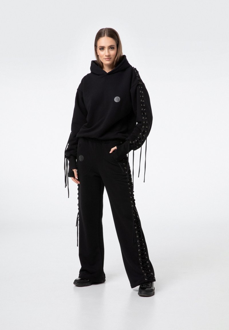 Czarny komplet z kapturem i spodniami, z ozdobnymi sznurowaniami na rękawach i bokach. Gładki materiał, luźny krój i okrągłe logo na bluzie.