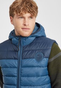 Pufferväst i mörkblått och marin med horisontell quiltning, dragkedjestängning och en rund logotyp på bröstet. Försedd med en huva.