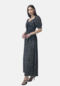 Robe maxi noire avec motif floral blanc, manches bouffantes, corsage ajusté et fente sur le côté, associée à des talons hauts noirs à lanières.