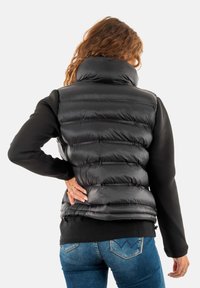 Gilet en duvet noir avec col haut, design matelassé et texture brillante. Porté sur une chemise noire à manches longues, associé à un jean en denim.