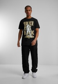 Sort bomulds t-shirt med et grafisk print af en fodboldspiller og teksten "TAKE THE KICK" i guld, parret med sorte cargo-bukser og hvide sneakers.
