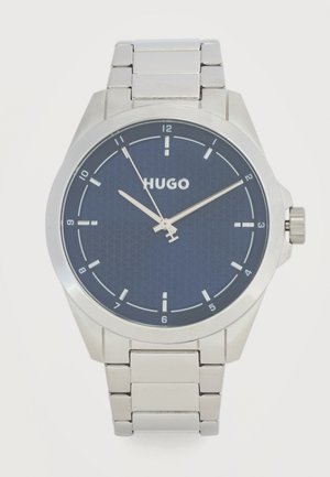 Sudraba metāla rokas pulkstenis ar zilu ciparnīcu, sudraba stundu marķieriem, trim rādītājiem un "HUGO" logotipu centrā.