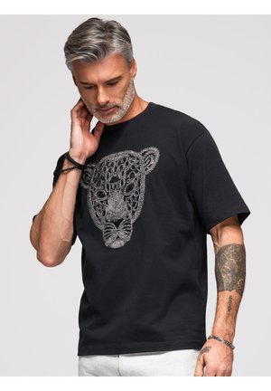 Homme aux cheveux gris et à la barbe portant un t-shirt noir avec un design complexe de visage de léopard argenté, touchant son cou et regardant vers le bas.