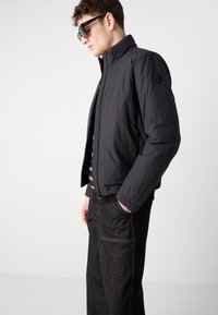 Jachetă bomber neagră cu un finisaj ușor texturat; guler în picioare și buzunare laterale; purtată peste o cămașă cu dungi și pantaloni din denim închis.