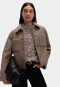 Braune Wolljacke mit Kragen, zwei Fronttaschen, Knopfverschluss, getragen über einem schwarz-braun gemusterten Hemd und kombiniert mit einer schwarzen Tasche.