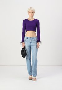 Violet cropped ilgas rankovėmis bluzonas su plunksnų akcentais ant rankovių, šviesiai mėlynos tiesios kojų džinsai ir juoda rankinė. Minimalistinis fonas.