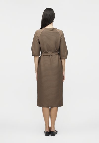 Γυναίκα με μακριά σκούρα μαλλιά φορά καφέ, με ζώνη, φόρεμα μήκους μέχρι τα γόνατα με μανίκια μέχρι τον αγκώνα, στέκεται με την πλάτη προς τον θεατή.