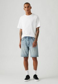 Weißes T-Shirt mit kurzen Ärmeln, hellblaue Jeansshorts mit ausgewaschenem Look und schwarze Turnschuhe. Weiße Kniestrümpfe sichtbar über den Schuhen.