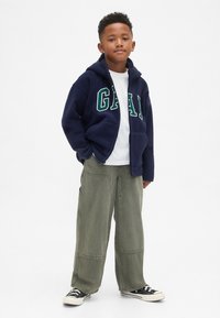 Laivastonsininen fleecetakki, jossa valkoiset ja vihreät "GAP"-kirjaimet, yhdistetty oliivinvihreisiin leveälahkeisiin housuihin ja mustiin kangaskenkiin.