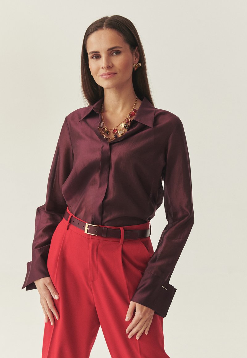 Donna con lunghi capelli castani che indossa una camicia di raso color bordeaux, pantaloni rossi, collana d'oro e rossa e orecchini d'oro, in piedi davanti a uno sfondo chiaro.