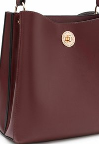 Bordeaux leren handtas met een zachte textuur, voorzien van een ronde gouden merkaccent en een gestructureerd, open ontwerp.
