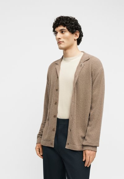 Άνδρας με σγουρά μαλλιά που φοράει μπεζ πλεκτή ζακέτα με κουμπιά πάνω από κρεμ μπλουζάκι και σκουρόχρωμο παντελόνι, κοιτάζοντας πλάγια σε λευκό φόντο.