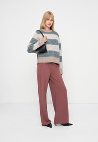 Mulher em pé, vestindo um suéter de malha listrado, calças largas cor malva, sapatos pretos de bico fino e carregando uma pequena bolsa preta no ombro.