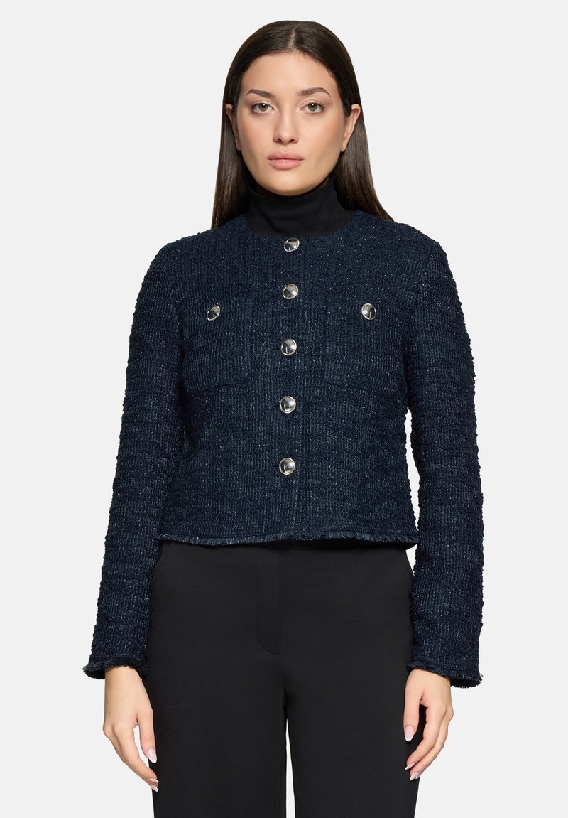 Femme aux cheveux bruns raides portant une veste boutonnée texturée bleu marine sur un col roulé noir et un pantalon noir, sur un fond uni.