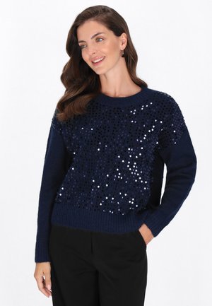 Femeie cu păr ondulat maroniu poartă un pulover bleumarin cu paiete strălucitoare și pantaloni negri, zâmbind cu mâna în buzunar, pe fundal alb.