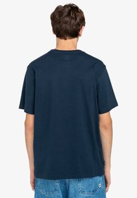 Camiseta de color azul marino de algodón, de manga corta, con cuello redondo y corte relajado. La vista trasera no muestra logotipos ni patrones visibles. Se lleva con pantalones cortos de mezclilla de color azul claro.
