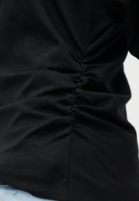 Tissu noir avec un détail de fronces réunies le long d'une couture verticale, porté sur du denim bleu clair au bord inférieur.