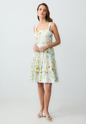 Witte zomerjurk met bloemen- en citroenpatronen, vierkante halslijn, brede bandjes, gelaagde rok, gemaakt van lichtgewicht stof, gecombineerd met witte sandalen.