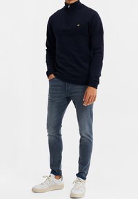 Suéter azul marino con cremallera, con un yugo texturizado, detalles acanalados y un logo dorado. Combinado con jeans azules y zapatillas blancas con detalles.