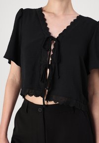 Top preto cropped com mangas curtas, acabamento em renda ondulada e fecho de laço na frente. Tecido macio e leve com um corte descontraído.