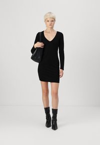 Vestido preto de mangas longas com decote em V e tecido texturizado. Modelo usa botas pretas de cano alto e folgadas, e segura uma mala preta.