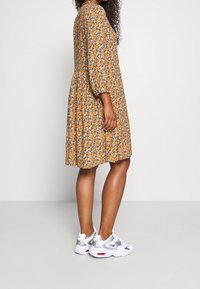 Blomstret knælængde kjole i sennepsgul med orange og hvide mønstre; har 3/4 ærmer og en indsnævret talje. Stylet med hvide sneakers.